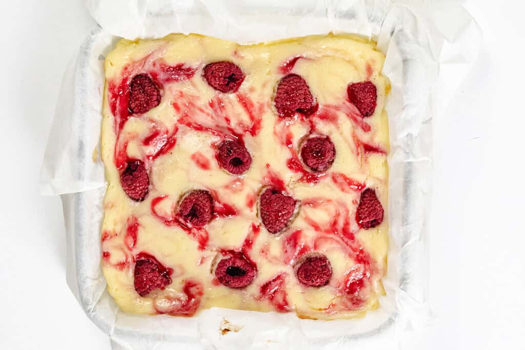 A square pan lined with parchment paper holds unbaked Raspberry Cheesecake Blondies batter, swirled with raspberry sauce and topped with whole raspberries.