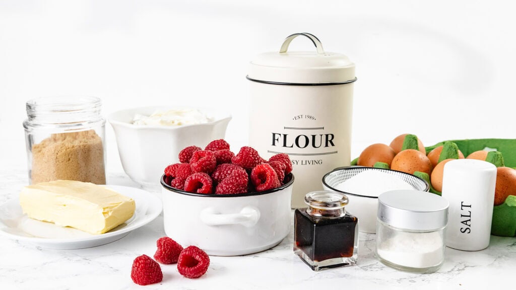 Baking ingredients on a white surface, including fresh raspberries in a bowl, butter, sugars, eggs in a carton, a flour canister, vanilla extract, cream, and salt—perfect for making delicious Raspberry cheesecake blondies.