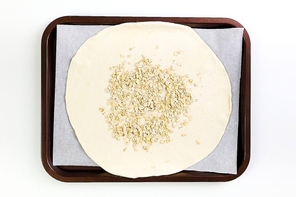 A rustic round sheet of dough topped with raw oats sits on parchment paper on a dark baking tray, ready to be transformed into a delicious summer Raspberry Galette.