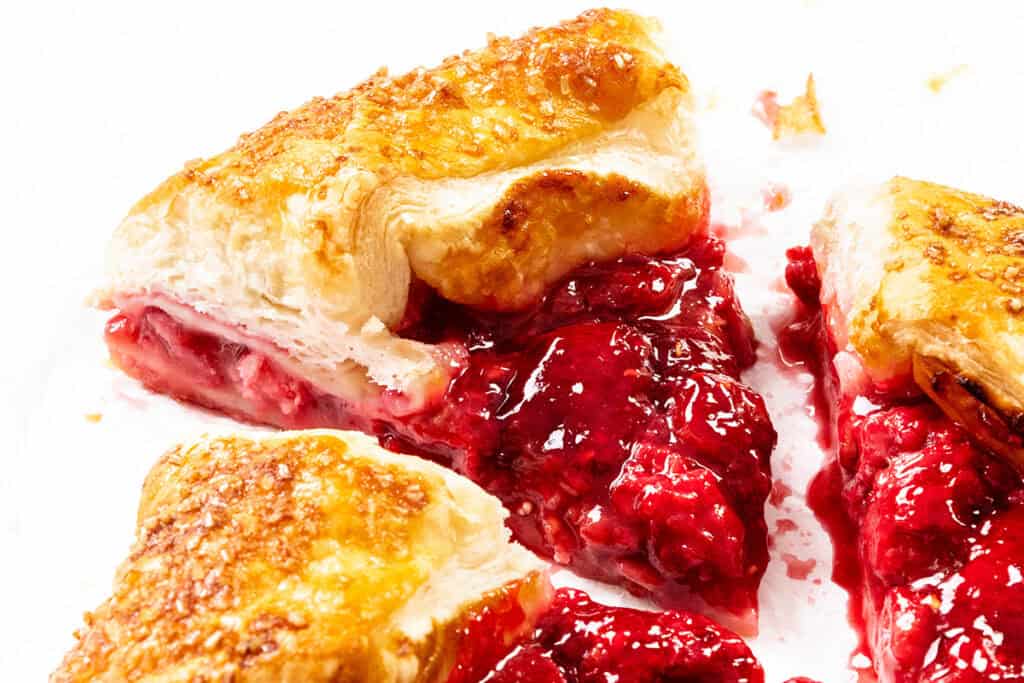 Close-up of a rustic galette, with flaky golden pastry slices filled with bright summer raspberry filling, bits of sugar on the crust, and some vibrant filling oozing out onto a white surface.