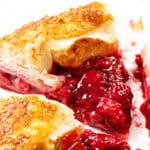 Close-up of a rustic galette, with flaky golden pastry slices filled with bright summer raspberry filling, bits of sugar on the crust, and some vibrant filling oozing out onto a white surface.