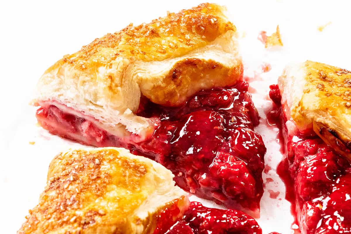 Close-up of a rustic galette, with flaky golden pastry slices filled with bright summer raspberry filling, bits of sugar on the crust, and some vibrant filling oozing out onto a white surface.