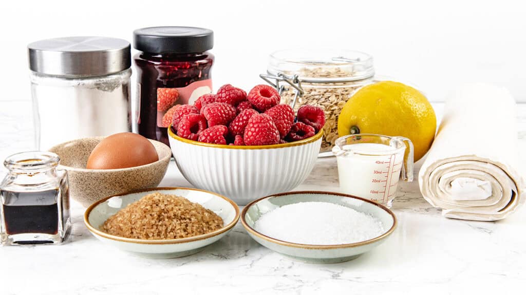 A bowl of fresh raspberries, a lemon, an egg, jars of sugar, jam, oats, vanilla extract, cream, and rolled pastry sheets arranged on a white countertop—perfect for making a rustic summer raspberry galette.