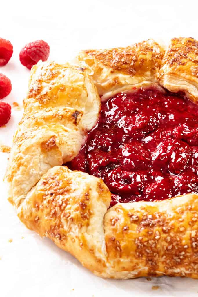 A close-up of a rustic summer raspberry galette with golden, flaky crust and glossy, red filling. Fresh raspberries are scattered in the background on a white surface.