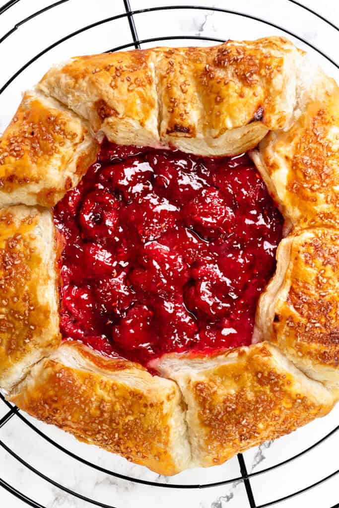 A close-up of a rustic summer raspberry galette with a golden, flaky crust and vibrant red raspberry filling, cooling on a black wire rack.