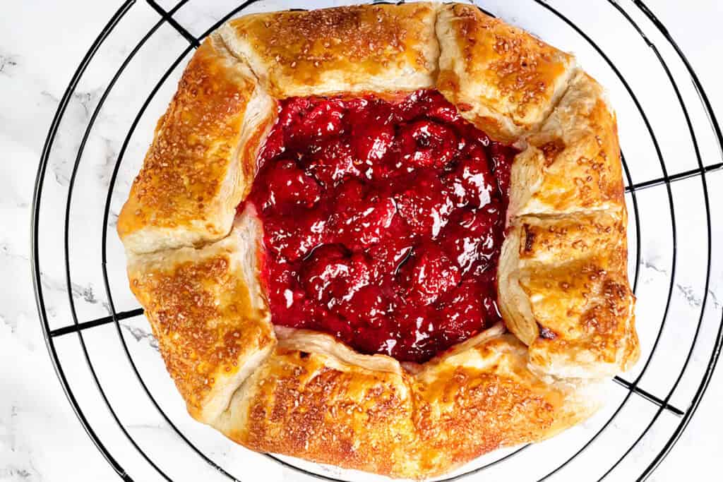 A rustic golden-brown raspberry galette with a flaky crust and a filling of bright red berries sits on a round cooling rack over a white marble surface—a perfect summer dessert.