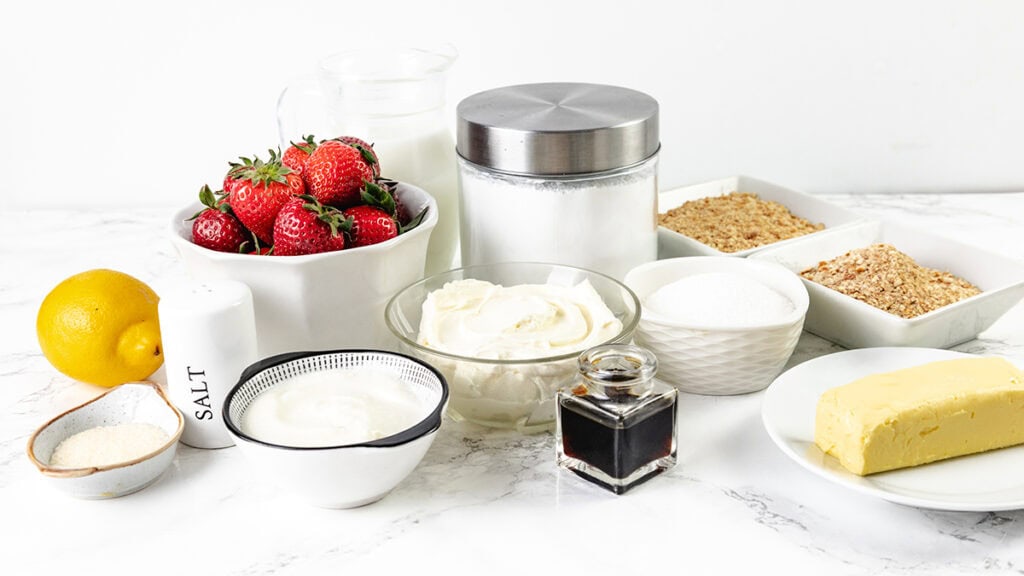 An assortment of homemade baking ingredients on a white surface, perfect for crafting a delicious dessert like Strawberry Pretzel Salad—featuring fresh strawberries, lemon, butter, sugar, vanilla extract, cream, yogurt, milk, and bowls of brown and white sugar.