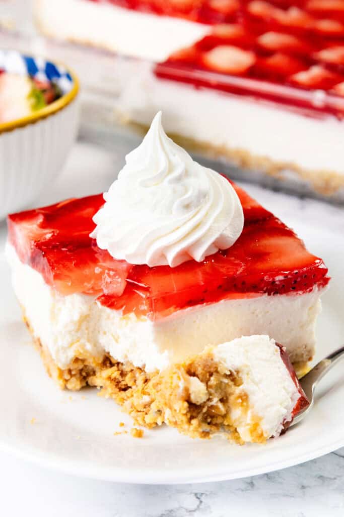 A slice of homemade Strawberry Pretzel Salad dessert with a pretzel crust, creamy filling, strawberry gelatin topping, and a swirl of whipped cream. A fork holds a bite on a white plate.