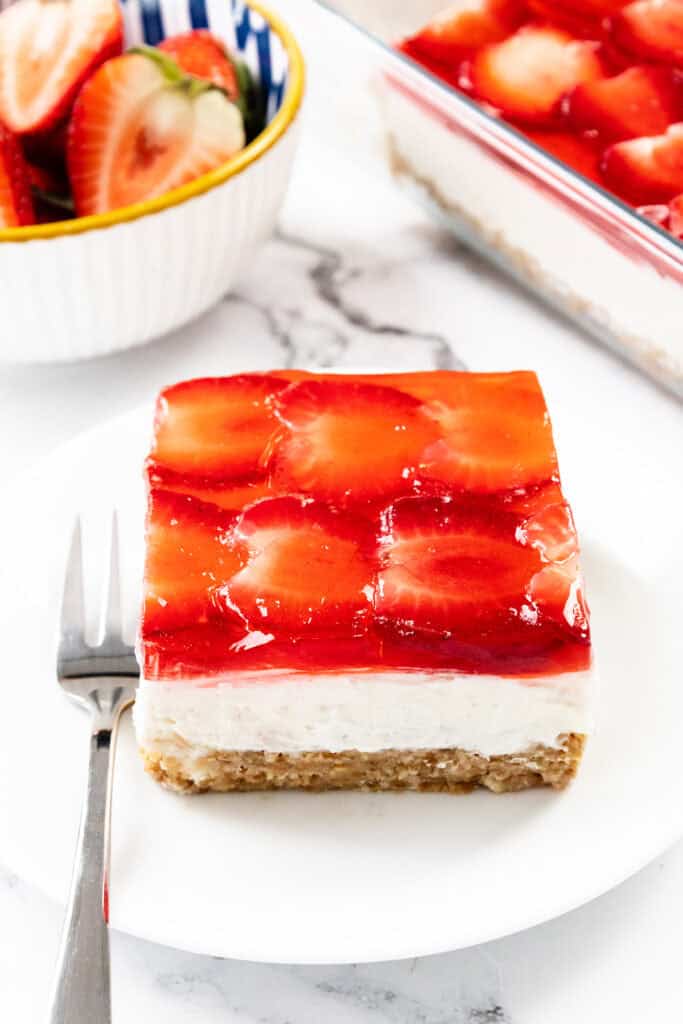A square slice of homemade Strawberry Pretzel Salad dessert, with a red gelatin and strawberry topping, creamy middle layer, and pretzel crust, sits on a white plate with a fork. A bowl of fresh strawberries and a baking dish are in the background.