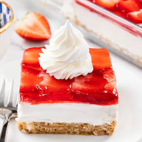 A slice of homemade Strawberry Pretzel Salad dessert with a crumbly crust, creamy middle layer, and strawberry topping, garnished with a swirl of whipped cream on a white plate, with a fork and fresh strawberries nearby.