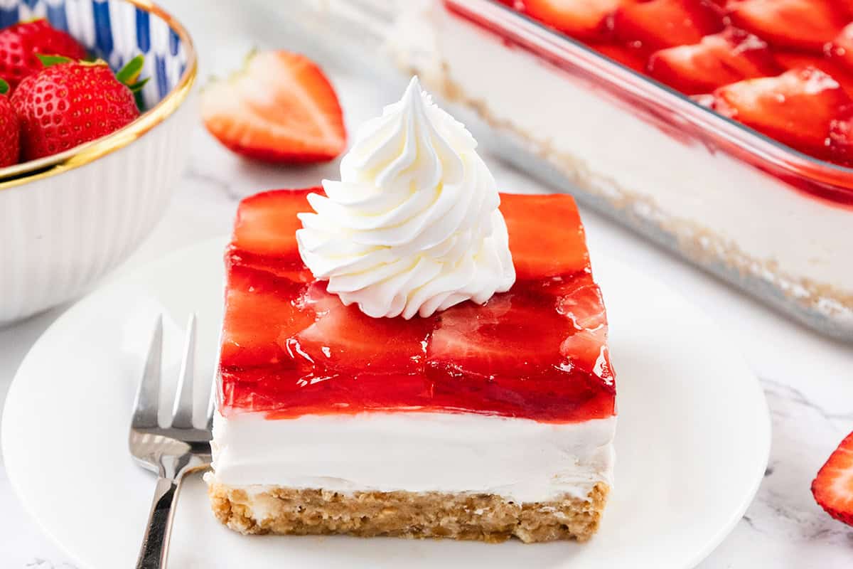 A slice of homemade Strawberry Pretzel Salad dessert with a crumbly crust, creamy middle layer, and strawberry topping, garnished with a swirl of whipped cream on a white plate, with a fork and fresh strawberries nearby.
