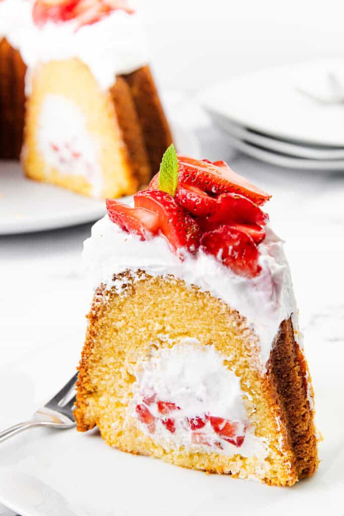 A slice of strawberry shortcake bundt cake, filled and topped with whipped cream and fresh strawberries, garnished with a mint leaf, sits on a white plate with a fork. The whole cake is in the background.