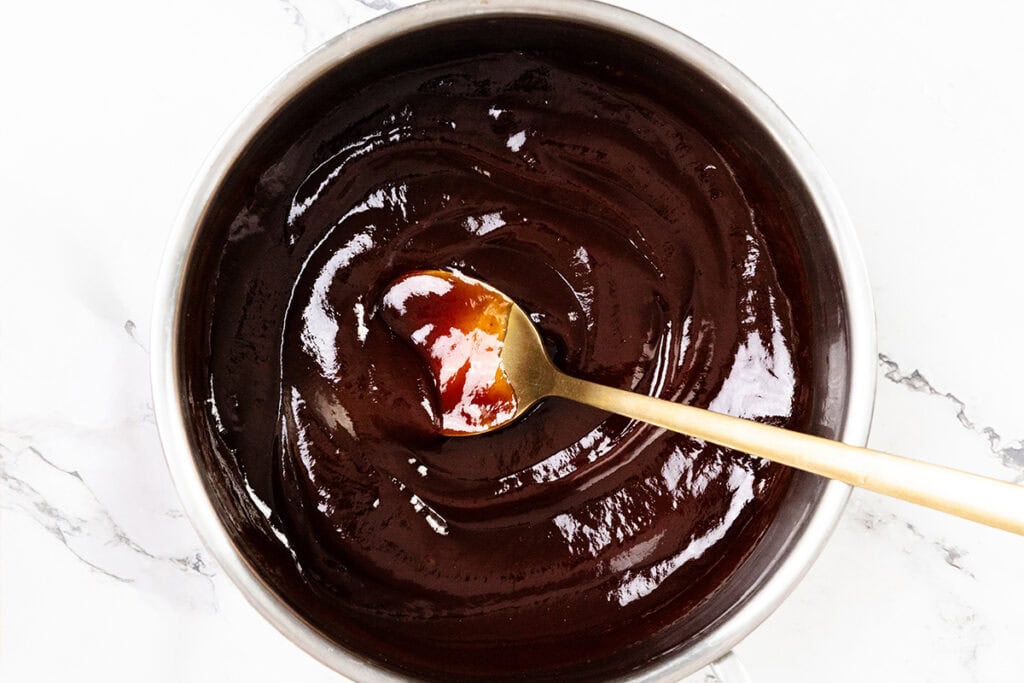 A saucepan filled with glossy, melted chocolate being stirred by a gold-colored spoon on a white marble surface.