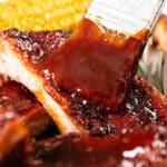 A close-up of barbecue ribs being brushed with a thick layer of sauce, with a piece of corn on the cob in the background.