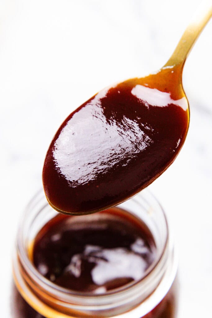 A close-up of a spoonful of thick, glossy barbecue sauce held over an open jar, with a white background.
