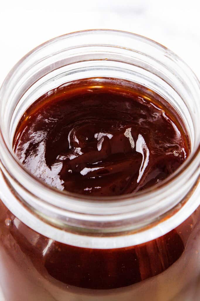 A close-up of an open glass jar filled with thick, glossy, dark brown barbecue sauce.