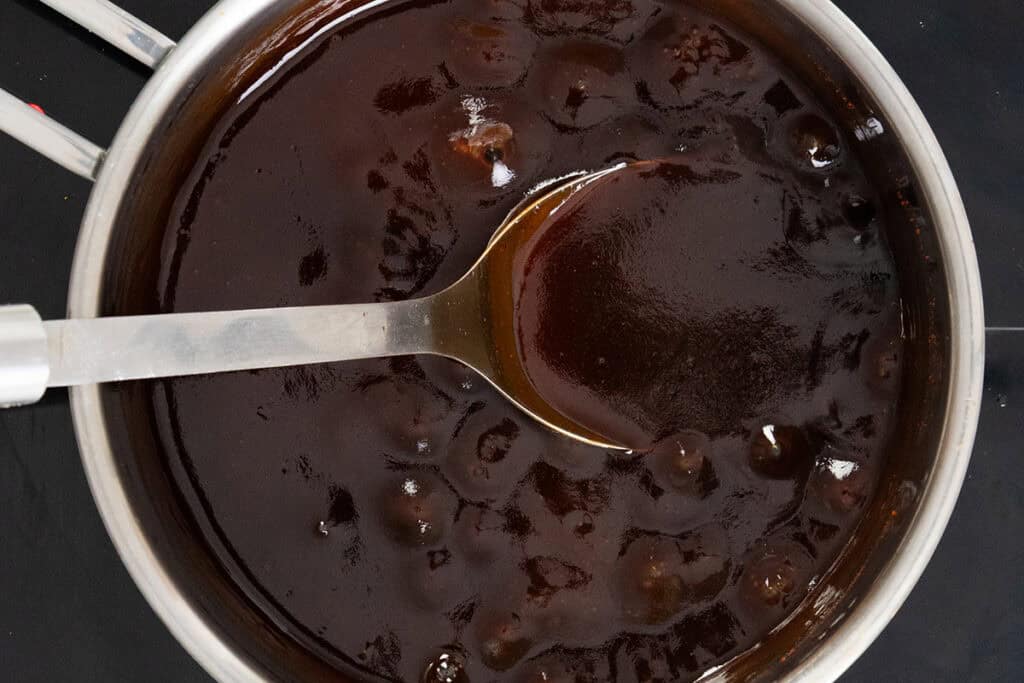 A metal pot filled with thick, dark brown sauce containing chunks, with a metal ladle resting inside, partially coated in the sauce.