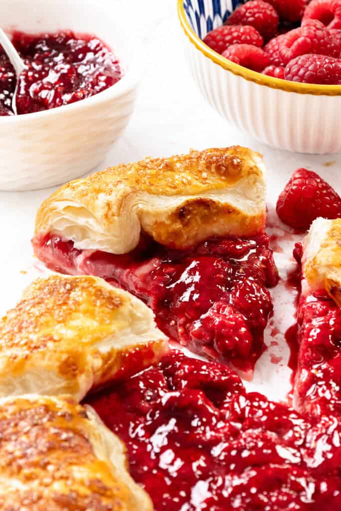 A close-up of a rustic raspberry galette, with flaky golden crust and oozing red fruit filling. Fresh raspberries and a bowl of raspberry jam sit in the background, evoking the flavors of summer.