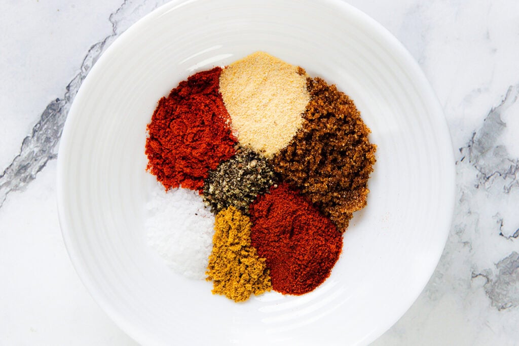 A white plate displays seven different ground spices arranged in separate piles, forming a colorful circular pattern on a marble surface.