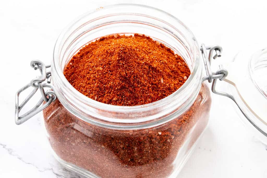 A clear glass jar filled with a reddish-brown spice blend, with the lid open, sitting on a white surface.