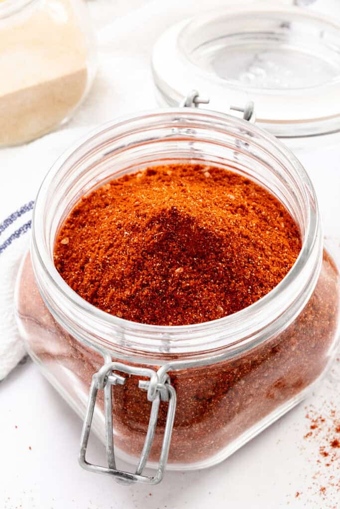 A glass jar filled with a reddish-brown spice blend sits open on a white surface, surrounded by a white cloth and another glass container in the background.