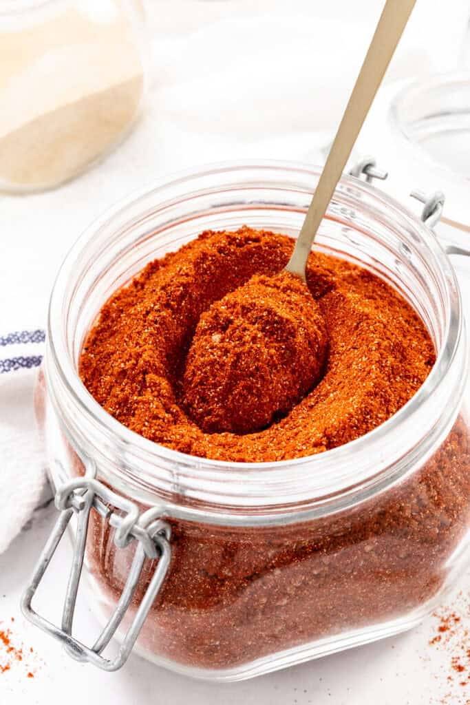 A glass jar filled with reddish-brown spice blend, with a metal spoon scooping some out. The jar has a hinged lid and sits on a white surface.