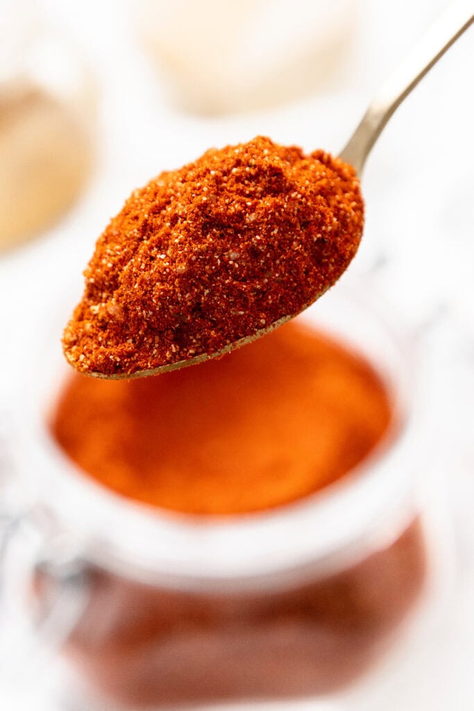 A close-up of a spoonful of reddish-brown spice blend held above an open glass jar filled with the same spice, against a softly blurred background.