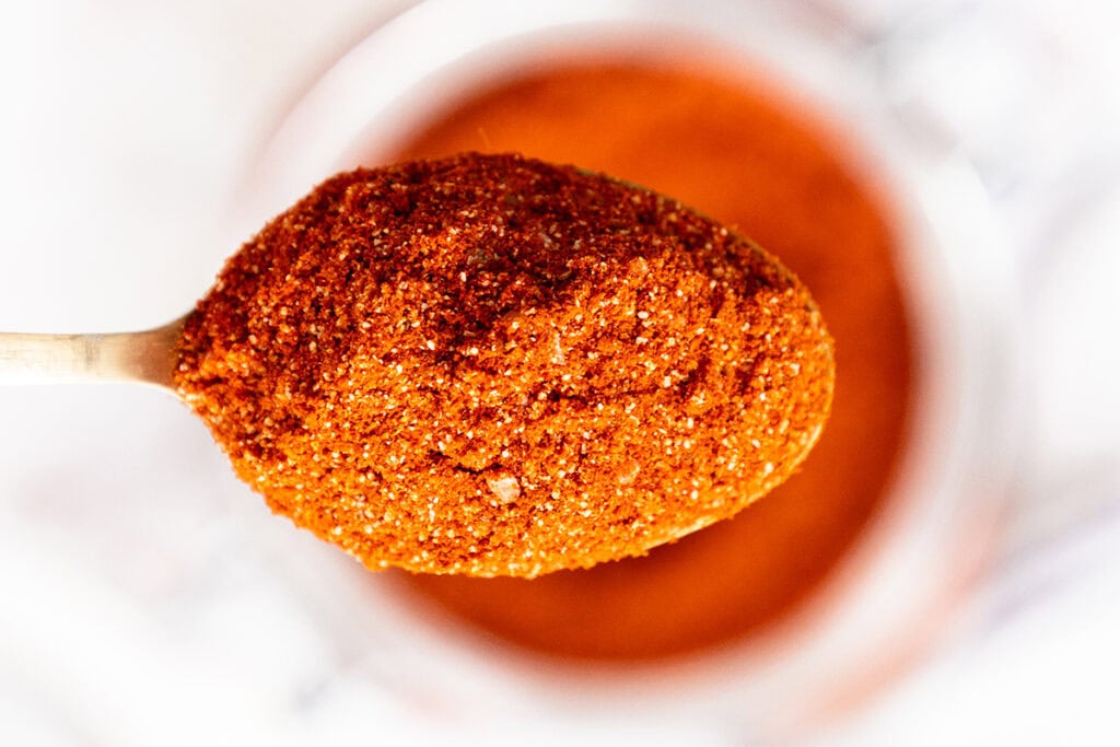 A close-up of a wooden spoon filled with a vibrant reddish-orange spice blend, held above a blurred container of the same spice in the background.