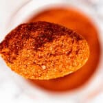 A close-up of a wooden spoon filled with a vibrant reddish-orange spice blend, held above a blurred container of the same spice in the background.