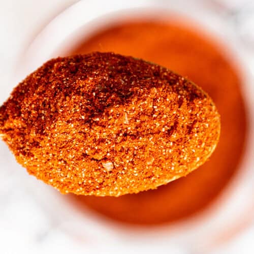 A close-up of a wooden spoon filled with a vibrant reddish-orange spice blend, held above a blurred container of the same spice in the background.