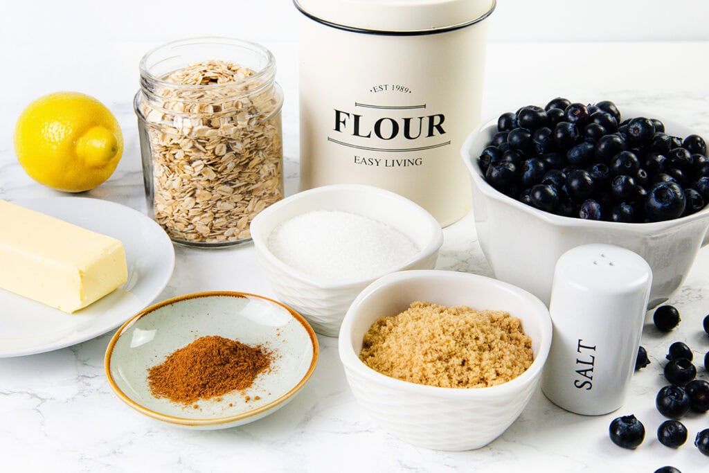 A display of baking ingredients for Blueberry Oatmeal Bars, including a stick of butter, a lemon, oats, flour, blueberries, white and brown sugar, cinnamon, and a salt shaker on a white countertop.