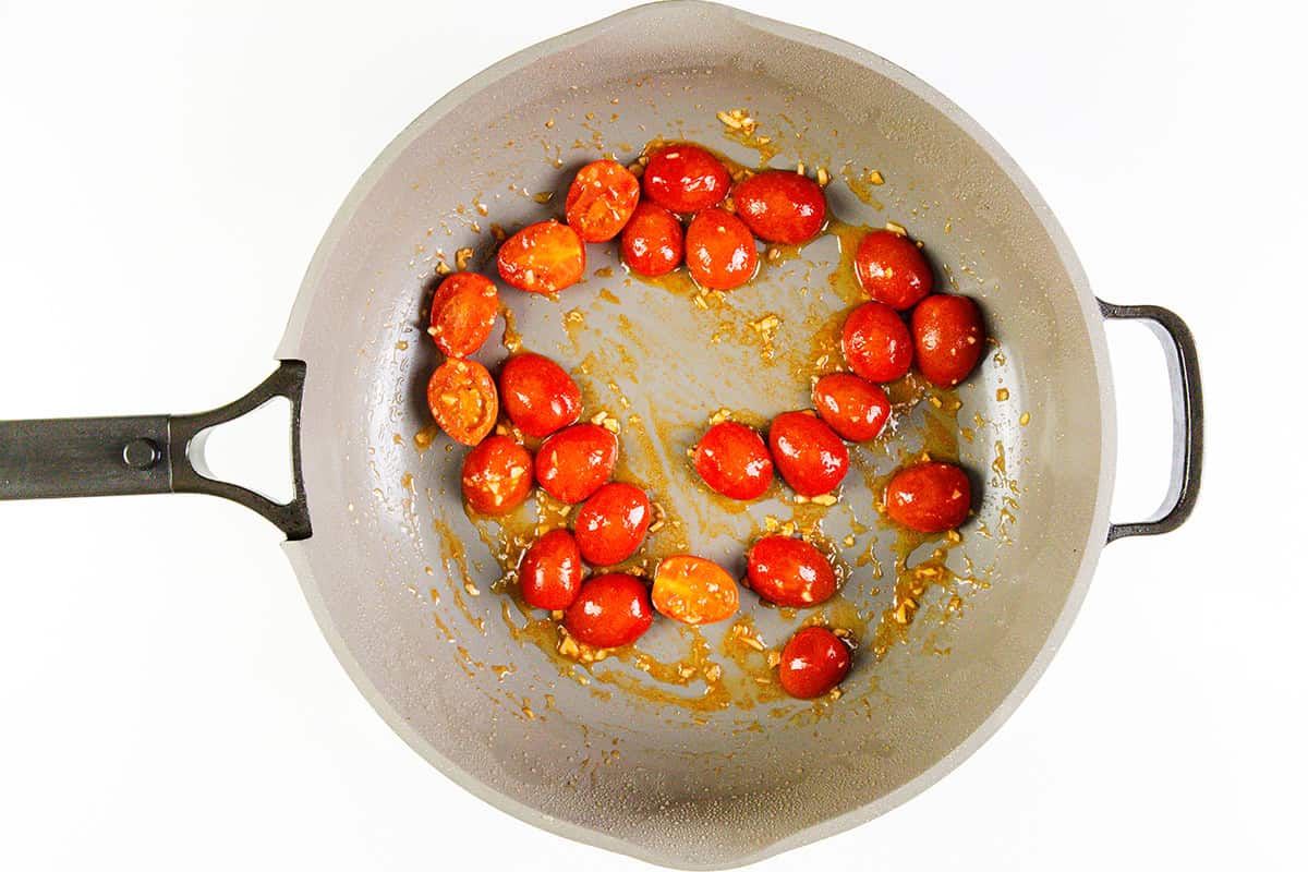 A grey skillet with sautéed garlic bits and halved cherry tomatoes arranged in a ring on the pan’s surface, perfect for starting Skillet Caprese Chicken, viewed from above on a white background.
