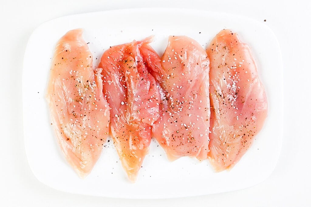 Four raw chicken breasts are arranged side by side on a white plate, sprinkled with coarse salt and black pepper—perfect for preparing Skillet Caprese Chicken. The background is white and clean.