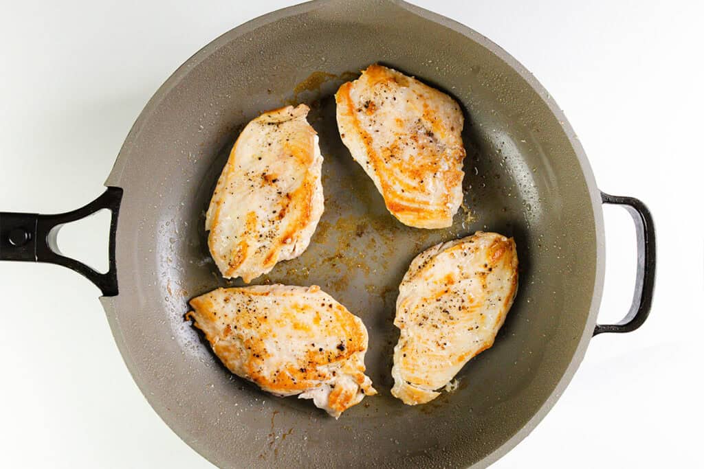 Four seasoned and cooked Caprese Chicken breasts are shown in a gray frying pan with a black handle, viewed from above on a white background.