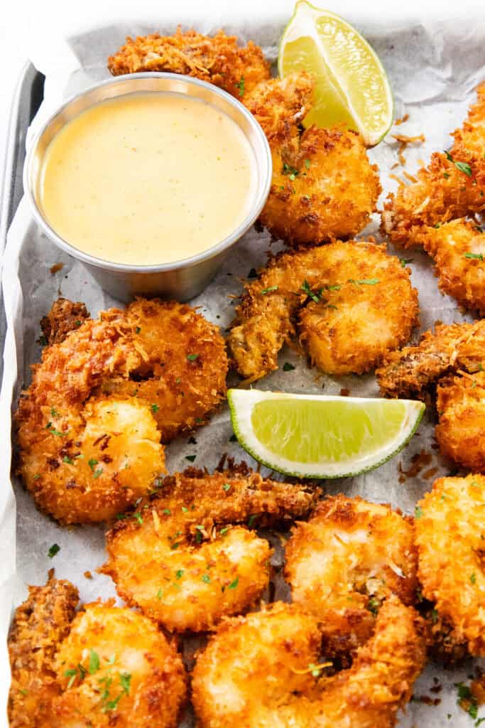 A tray of crispy coconut shrimp garnished with chopped herbs, served with lime wedges and a small metal cup of creamy dipping sauce on parchment paper.