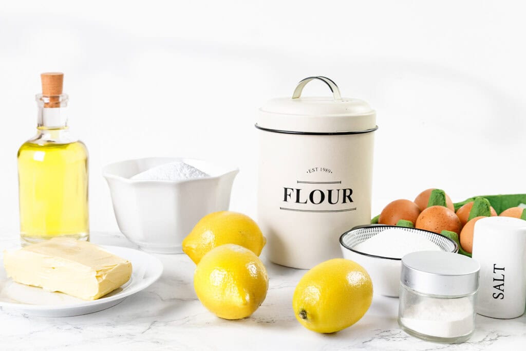 Baking ingredients for a Glazed Lemon Bundt Cake on a white surface: a bottle of oil, butter, three lemons, a flour canister, bowl of sugar, eggs, sieve, salt shaker, and a small jar of sugar.