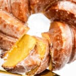 A close-up of a Glazed Lemon Bundt Cake on a glass plate with a golden knife cutting a slice. The cake features a shiny, sugary lemon glaze and a moist, yellow interior.