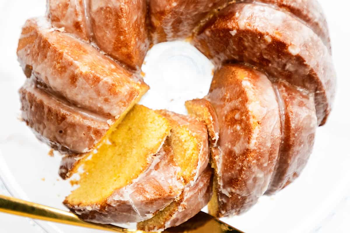 A close-up of a Glazed Lemon Bundt Cake on a glass plate with a golden knife cutting a slice. The cake features a shiny, sugary lemon glaze and a moist, yellow interior.