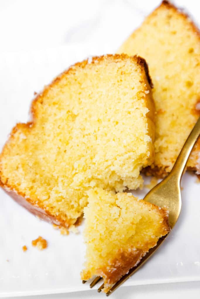 A close-up of two slices of Glazed Lemon Bundt Cake on a white plate, with a gold fork cutting a piece from one slice. The cake looks moist and has a slightly crumbly texture.