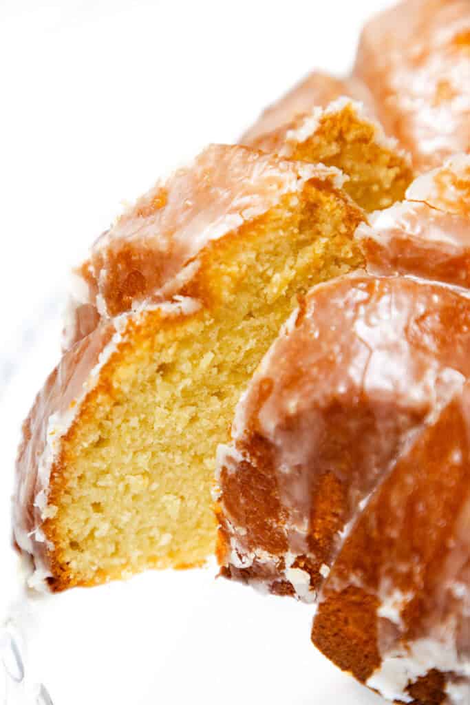 A close-up of a sliced Glazed Lemon Bundt Cake reveals a golden, moist interior and a shiny sugar glaze coating the outside.