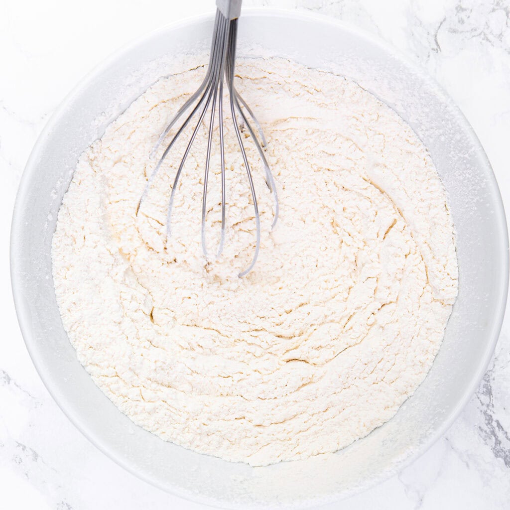 A metal whisk sits in a large white bowl filled with dry flour mixture for a Glazed Lemon Bundt Cake, set on a white marble surface.