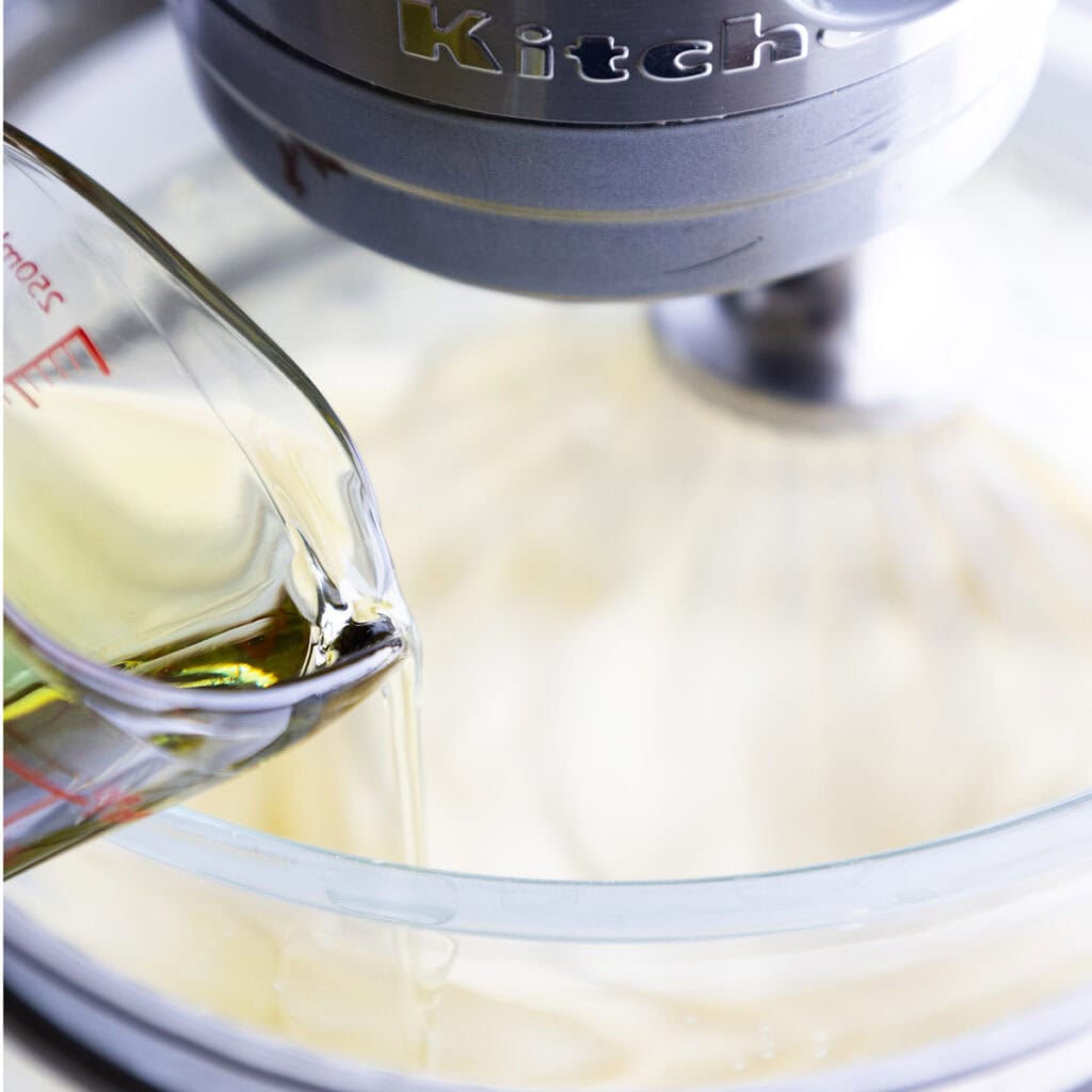 A close-up of oil being poured from a glass measuring cup into a stand mixer bowl with Lemon Bundt Cake batter inside. The mixer is running, and part of the metallic KitchenAid logo is visible.
