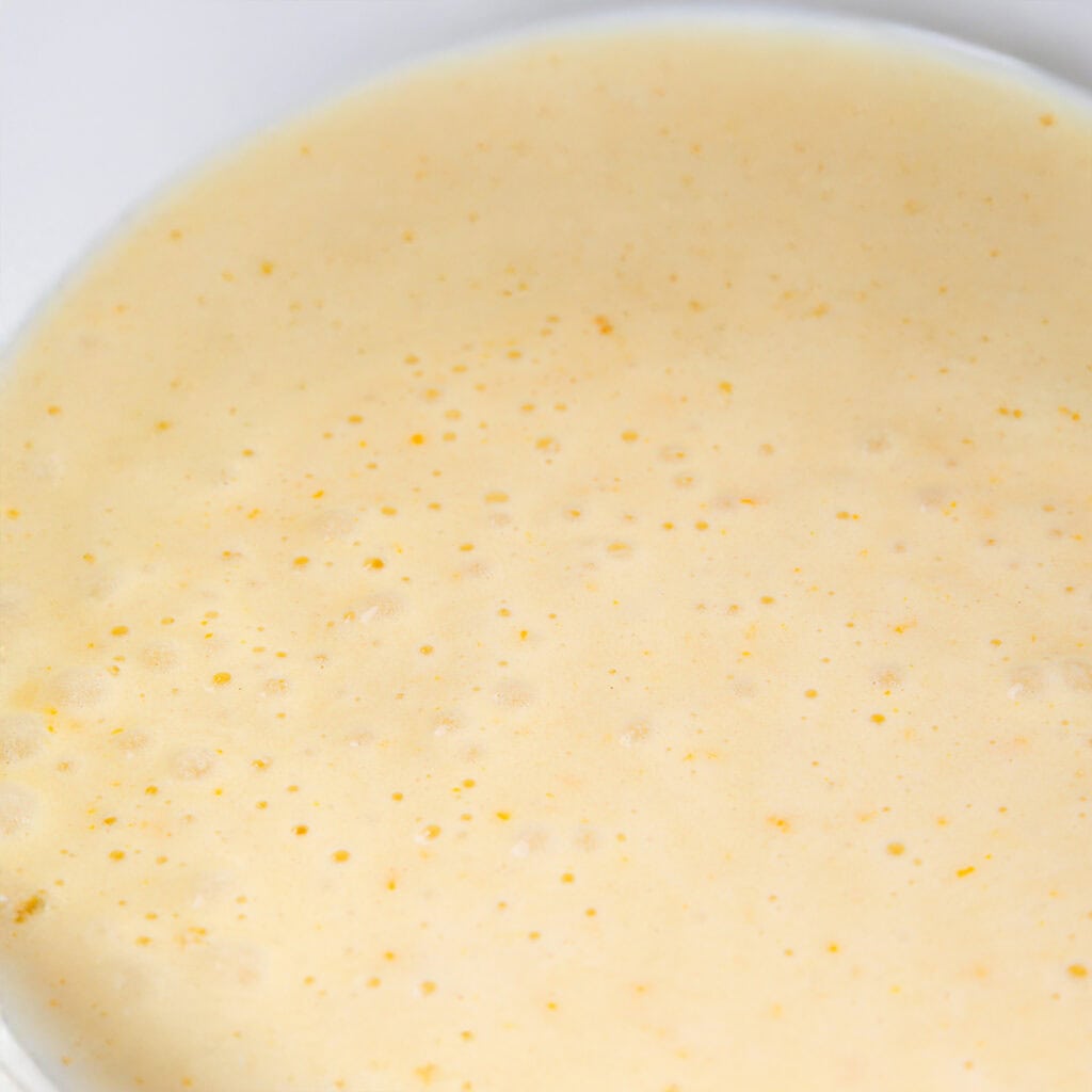 Close-up of a creamy, pale yellow sauce with small bubbles and specks on its surface, suggesting the smooth, slightly thick texture perfect for drizzling over a Lemon Bundt Cake.