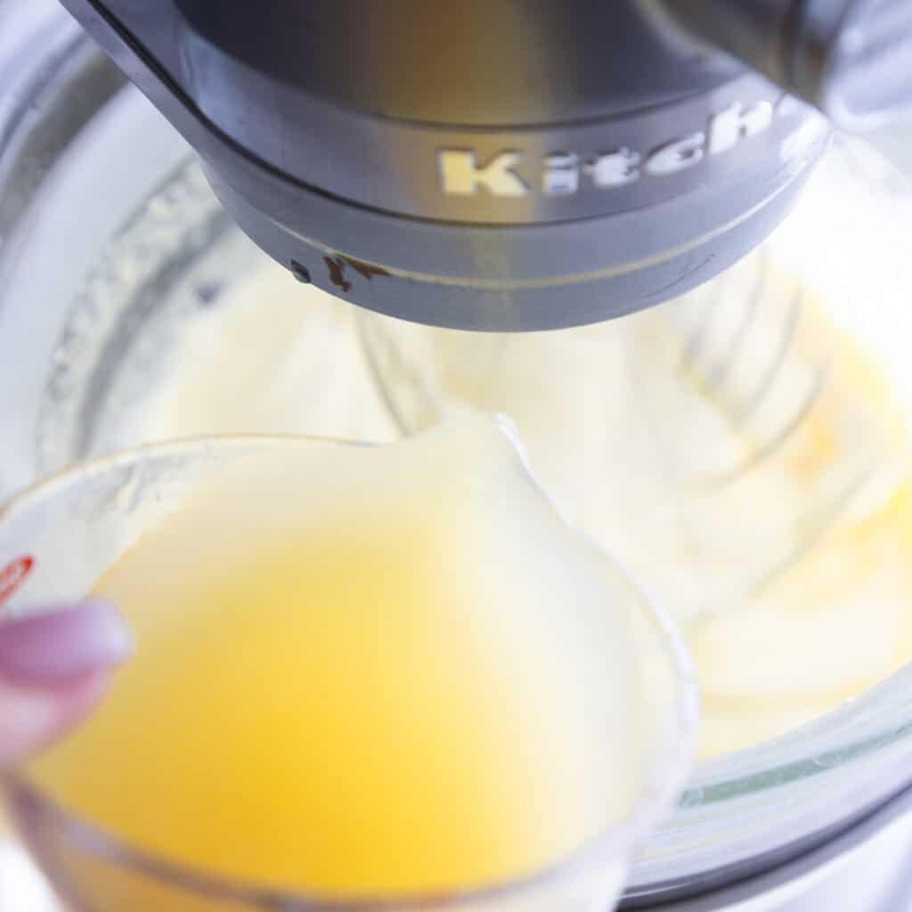 A hand pours a yellow liquid from a measuring cup into a stand mixer with a glass bowl, preparing ingredients for a Lemon Bundt Cake. The mixer is branded “KitchenAid.”.