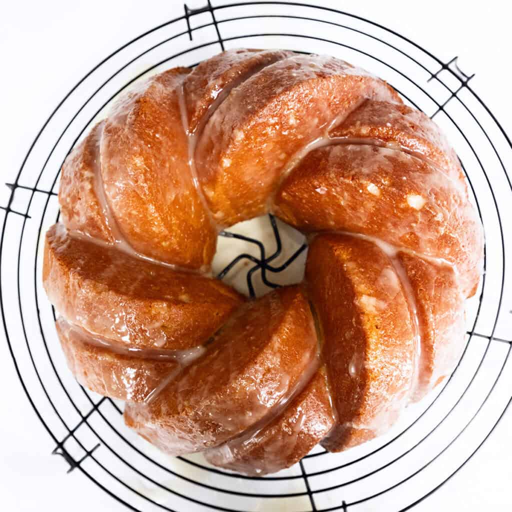 A Glazed Lemon Bundt Cake, golden brown and topped with a light glaze, sits on a round black wire cooling rack, viewed from above against a white background.