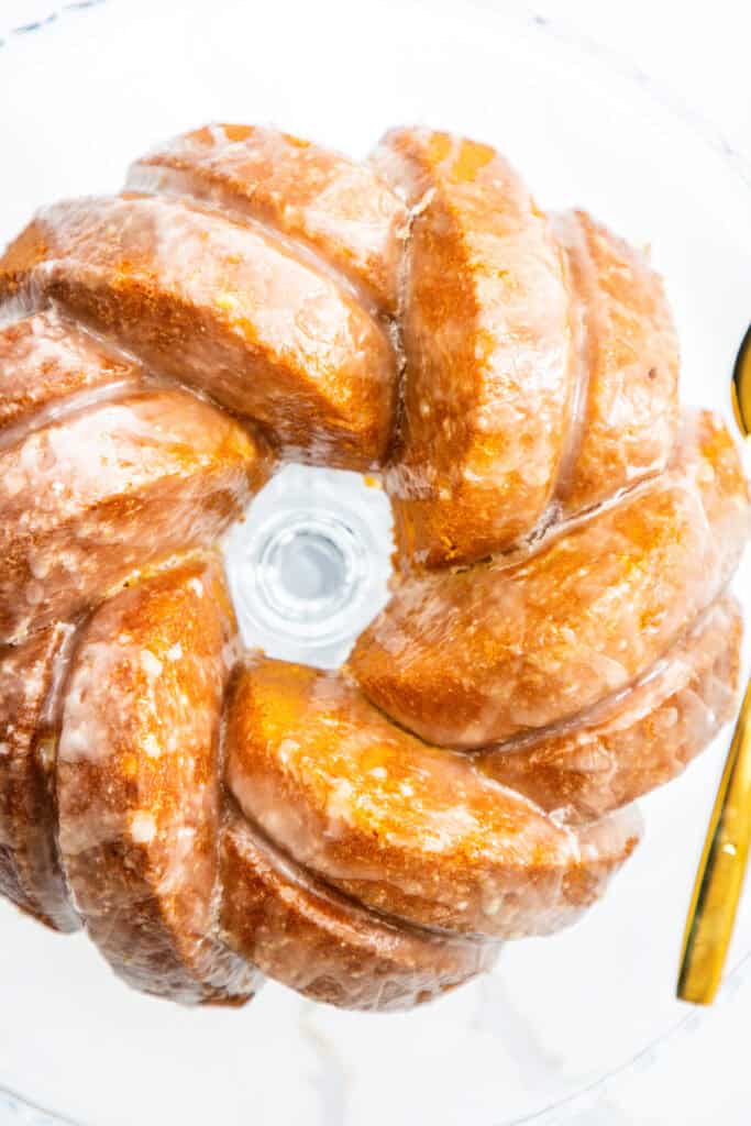 A Glazed Lemon Bundt Cake with a swirled design sits on a glass cake stand, viewed from above. The cake features a shiny, sugary coating that enhances its appeal.