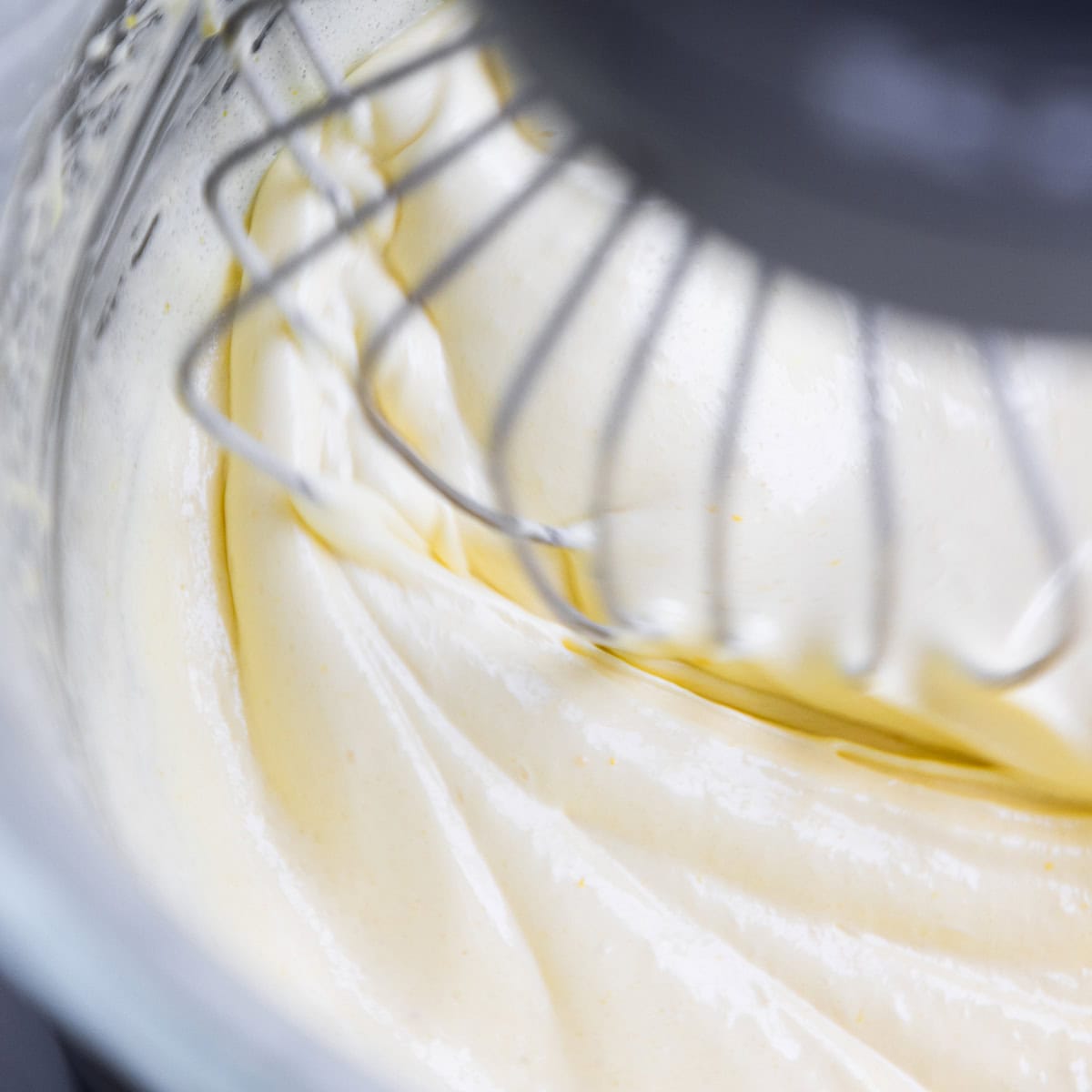 Close-up of a metal whisk mixing creamy, light yellow batter in a glass bowl, creating smooth, swirling patterns—the start of a delicious Glazed Lemon Bundt Cake.