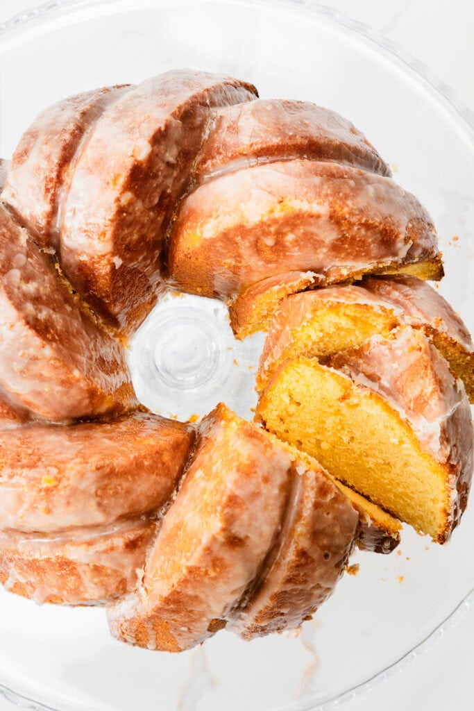 A glazed lemon bundt cake with a golden yellow interior sits on a glass cake stand. Several slices have been cut and slightly separated, revealing the moist texture inside.