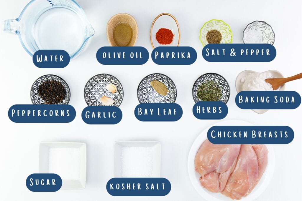 Top-down view of labeled brined chicken ingredients, including water, olive oil, paprika, salt, peppercorns, garlic, bay leaf, herbs, baking soda, sugar & kosher salt—everything you need for perfect grilled chicken breast in separate bowls.
