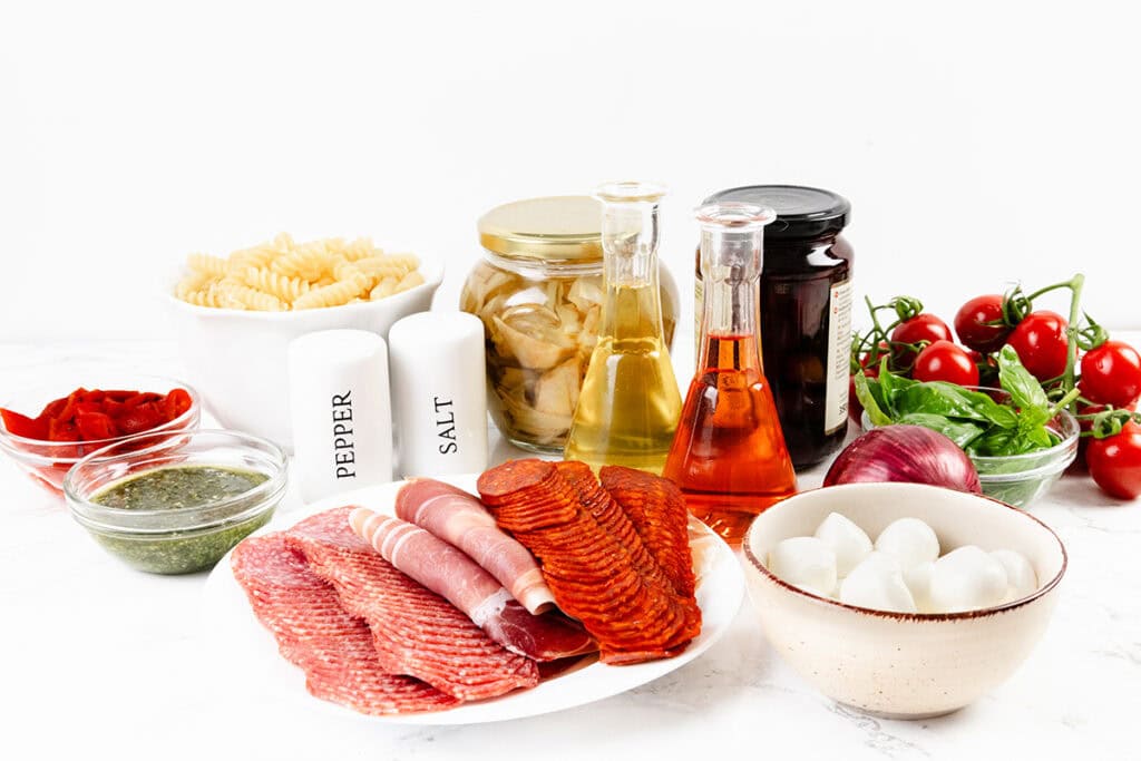 A selection of Italian antipasto ingredients on a white surface, including cured meats, mozzarella balls, pasta for easy recipes like pasta salad, jars of oil and vegetables, pesto, tomatoes, basil, red onion, and seasonings.